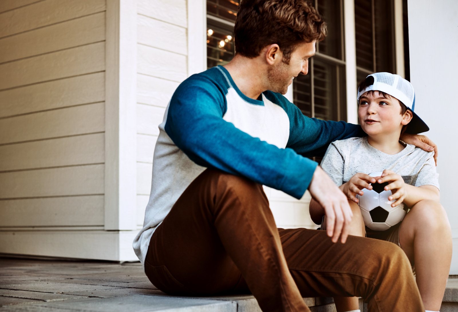 A father sits with his son on their front porch, talking and listening as the boy holds a soccer ball