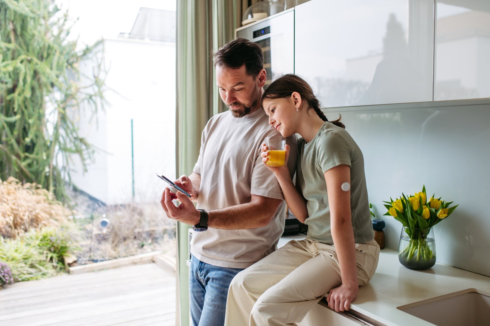 A father shows his daughter something on his phone while she drinks orange juice with him in the kitchen